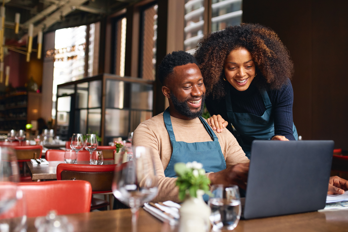 decorative image of a man and woman wearing aprons in a restaurant and standing behind a computer